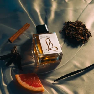 Louloudion TRACE 50 ml Eau de Parfum şişesi; etrafında taze greyfurt dilimi, tarçın çubukları, vanilya çubuğu ve kıyılmış tütün ile dökümlü kumaş üzerinde gösterimi.